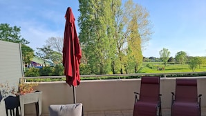 A balcony with two lounge chairs, a red umbrella, and a view of a park.