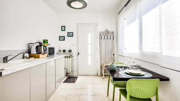 A modern kitchen with white cabinets, a dining table with green chairs, and a window with blinds.