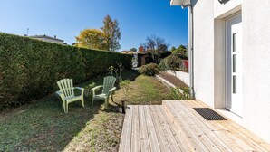 A wooden deck with two chairs, a hedge, and a white building.