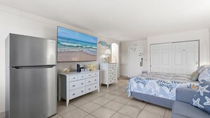 Quarto com cama, cômoda, geladeira e televisão exibindo uma cena de praia.
