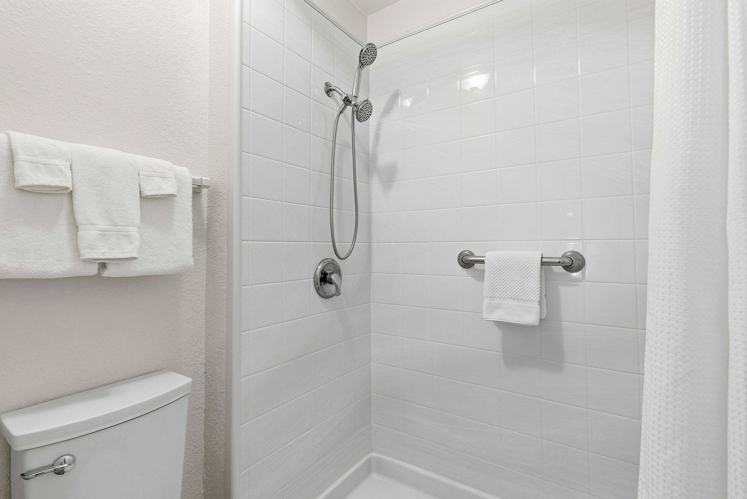 Een badkamer met witte betegelde wanden, een douche met handdouche en twee handdoekrekken met witte handdoeken.