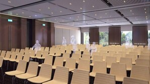 Ein Konferenzraum mit Reihen von Stühlen, einer modernen Decke mit eingelassener Beleuchtung und großen Fenstern mit Blick auf Grünflächen.