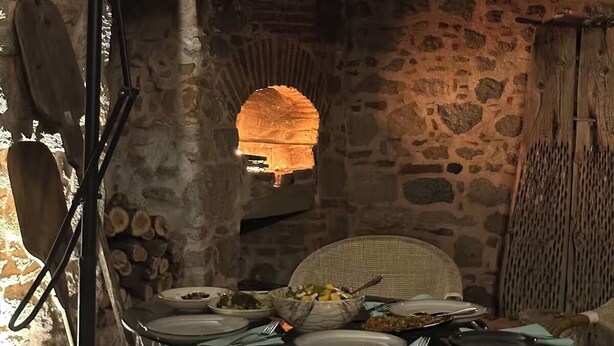 A rustic stone interior with a dining table set for a meal, surrounded by stone walls and a hanging light fixture.