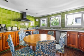 Una cocina con baldosas verdes, armarios de madera, una mesa redonda, sillas estampadas y un ventanal con vistas a la vegetación.