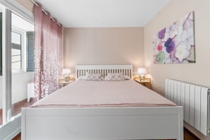 A bedroom with a bed, bedside lamps, a window with curtains, a radiator, and a floral wall art piece.