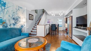 A modern living room with a blue sofa, a glass coffee table, a staircase, a dining area, and a kitchen with a TV. - Holiday Home 'Deti' with Private Terrace and Wi-Fi (Roses)
