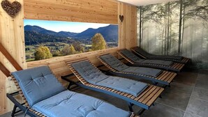 Una habitación de madera con cuatro sillones reclinables, un ventanal con vistas a la montaña y un mural de un bosque en la pared.