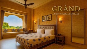 A spacious bedroom with a large bed, a view through a window, and a ceiling fan. - Hotel Grand Haveli Jaisalmer (Jaisalmer)