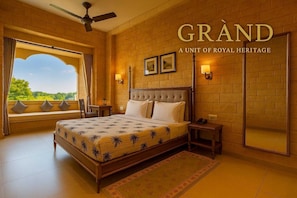 A spacious bedroom with a large bed, a view through a window, and a ceiling fan. - Hotel Grand Haveli Jaisalmer (Jaisalmer)