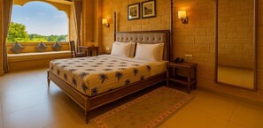 A spacious bedroom with a large bed, a view through a window, and a ceiling fan. - Hotel Grand Haveli Jaisalmer (Jaisalmer)
