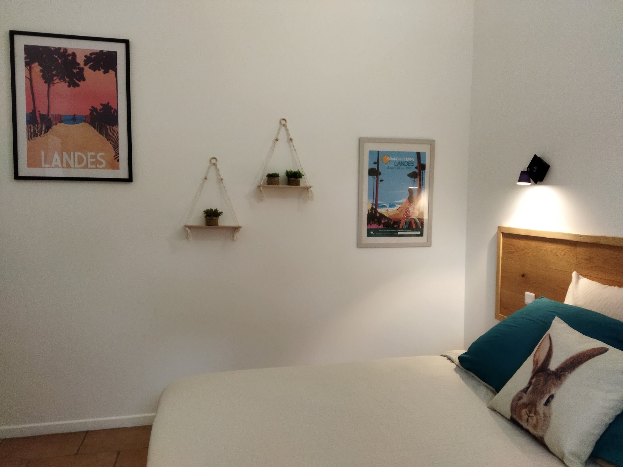 Une chambre à coucher comprenant un lit, une tête de lit, une étagère fixée au mur avec une plante, une affiche encadrée et un luminaire fixé au mur.