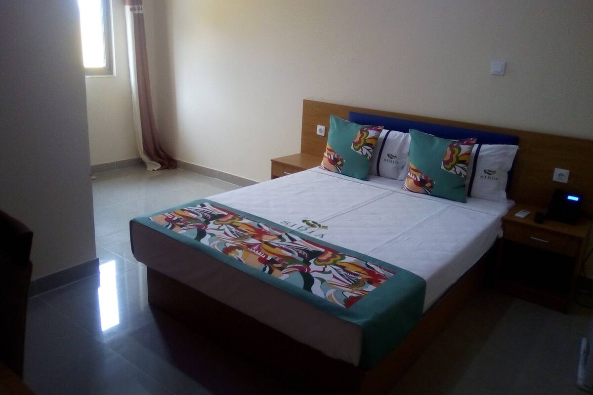 Une chambre d’hôtel avec un grand lit, une table de chevet, une commode et une fenêtre avec des rideaux.