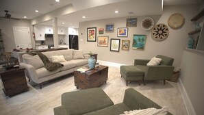 A modern living room with a sofa, a coffee table, and a green armchair. There is a kitchen in the background with white cabinets and a refrigerator. - Eclectic Charm (Douglasville)