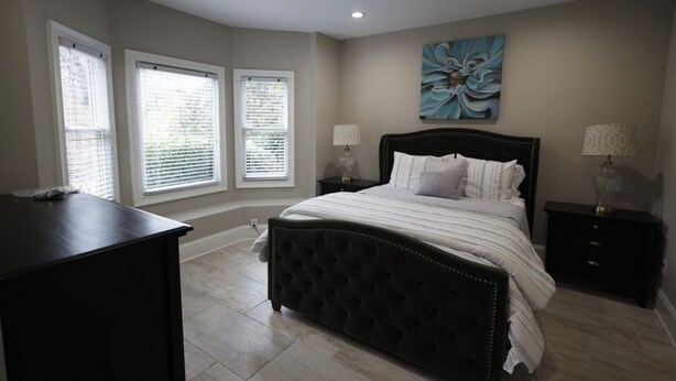 A bedroom with a large bed, two bedside tables with lamps, a dresser, and three windows with blinds. - Eclectic Charm (Douglasville)