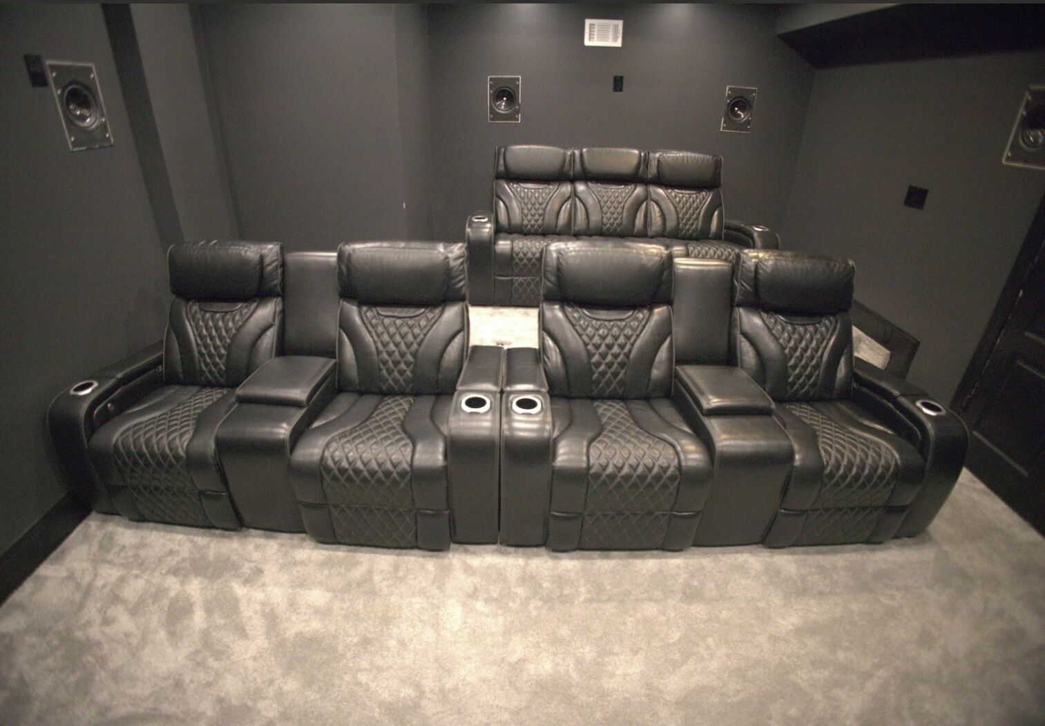 A home theater room with a curved black leather couch, a central console, and wall-mounted speakers.