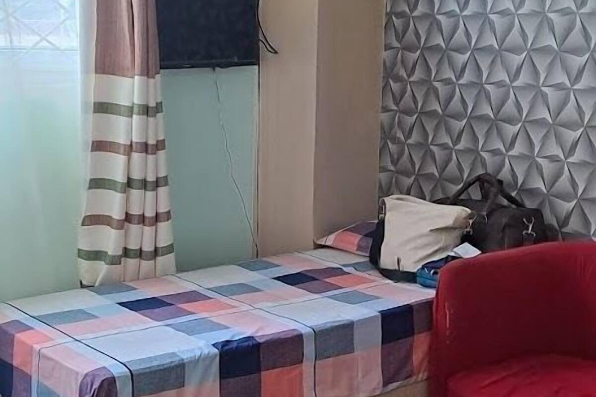Ένα υπνοδωμάτιο με ένα κρεβάτι, μια κόκκινη καρέκλα, μια τηλεόραση τοποθετημένη στον τοίχο και ένα παράθυρο με κουρτίνες.