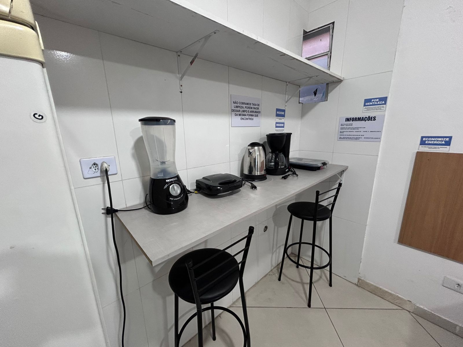 A kitchenette with a blender, coffee maker, and a small refrigerator. There are two black stools and a white countertop.