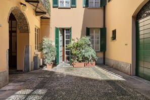 A narrow cobblestone alley with a building entrance, green shutters, and potted plants.