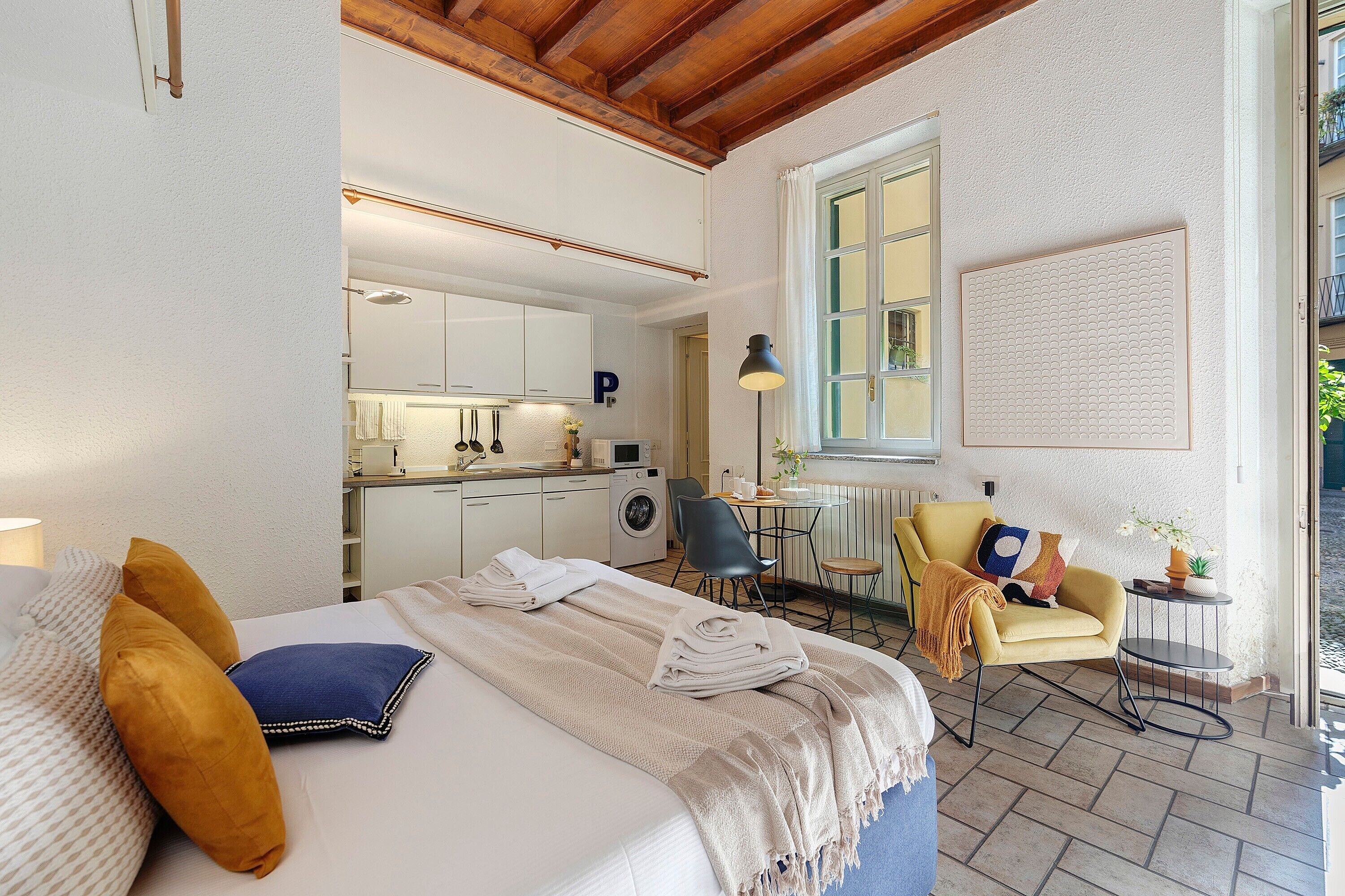 Un dormitorio con cama, almohadas, una mesita de noche, una silla, una pequeña mesa, una ventana y una zona de cocina al fondo.