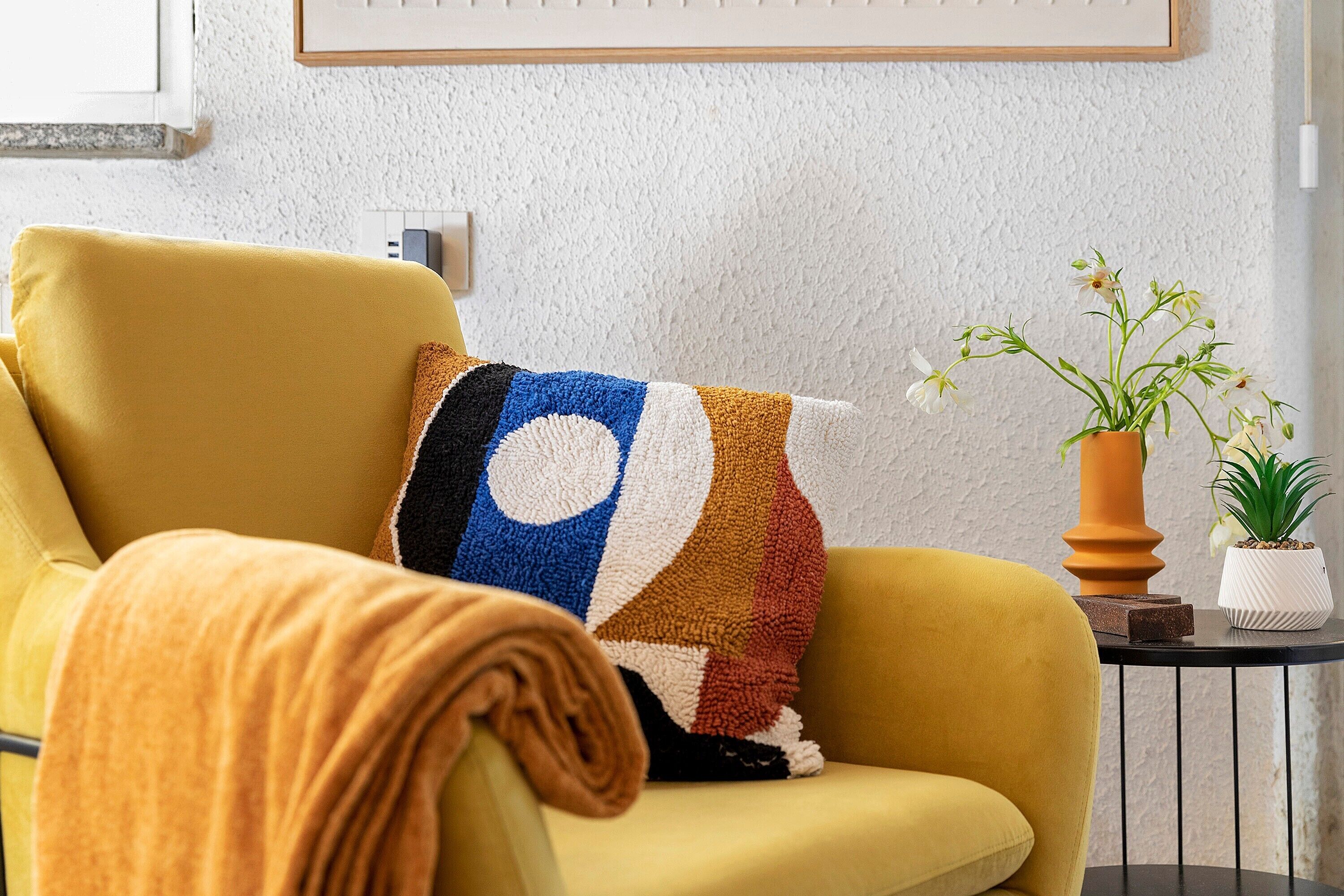 Un fauteuil jaune agrémenté d’un coussin aux couleurs vives, une petite table sur laquelle est posée une plante en pot et un tableau encadré accroché au mur.