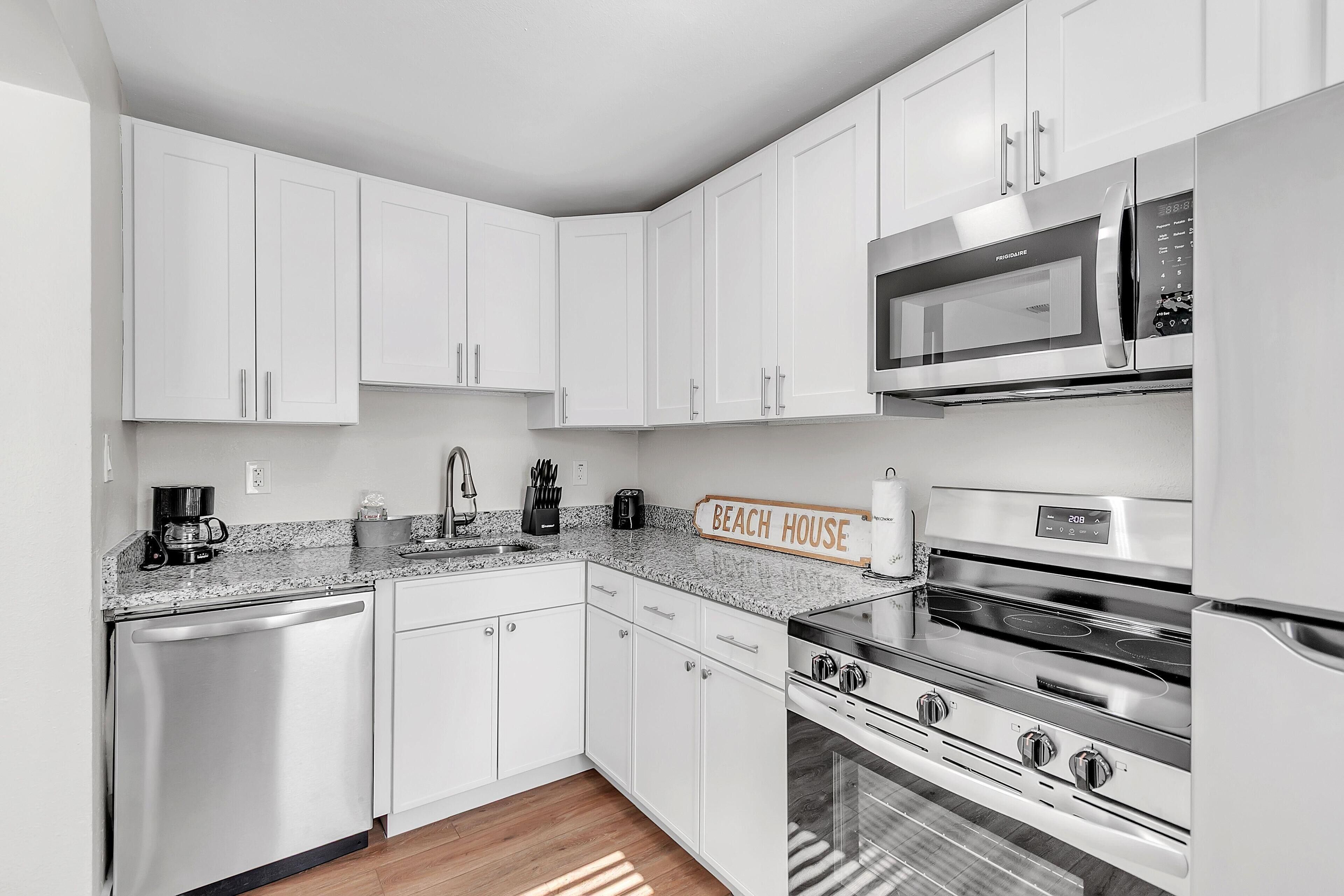 A modern kitchen with white cabinetry, a granite countertop, a stainless steel refrigerator, a microwave, a stove, and a coffee maker.