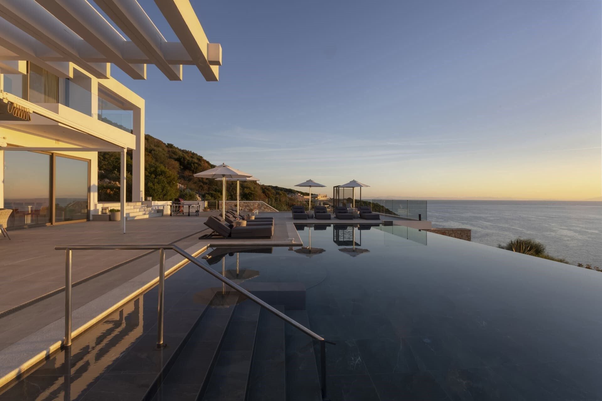A modern infinity pool with a glass edge overlooking the sea, outdoor seating, and a contemporary building with large windows.