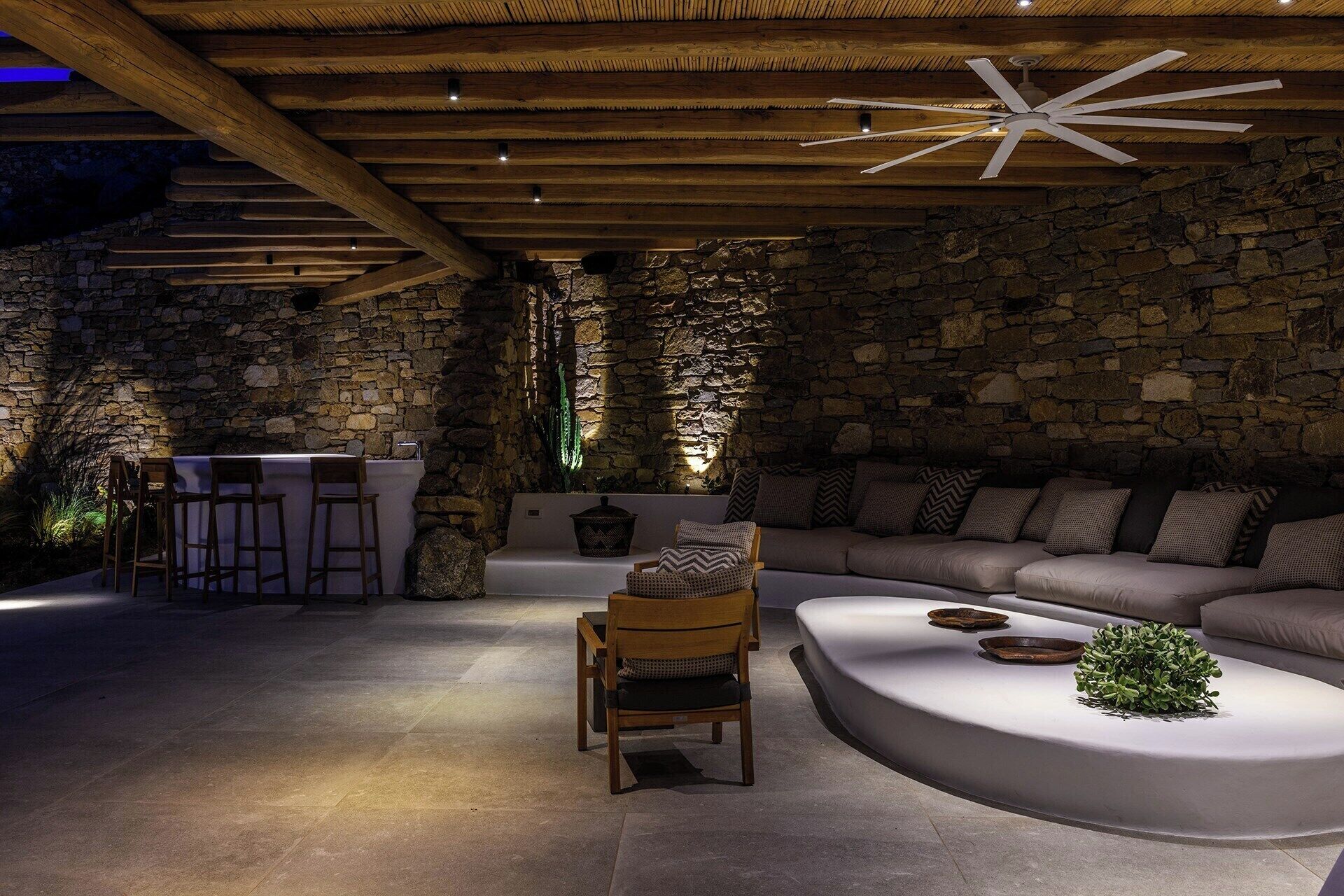 A cozy interior with a stone wall, wooden ceiling beams, a stone bench, a wooden chair, a white sofa, and a small plant on a white table.
