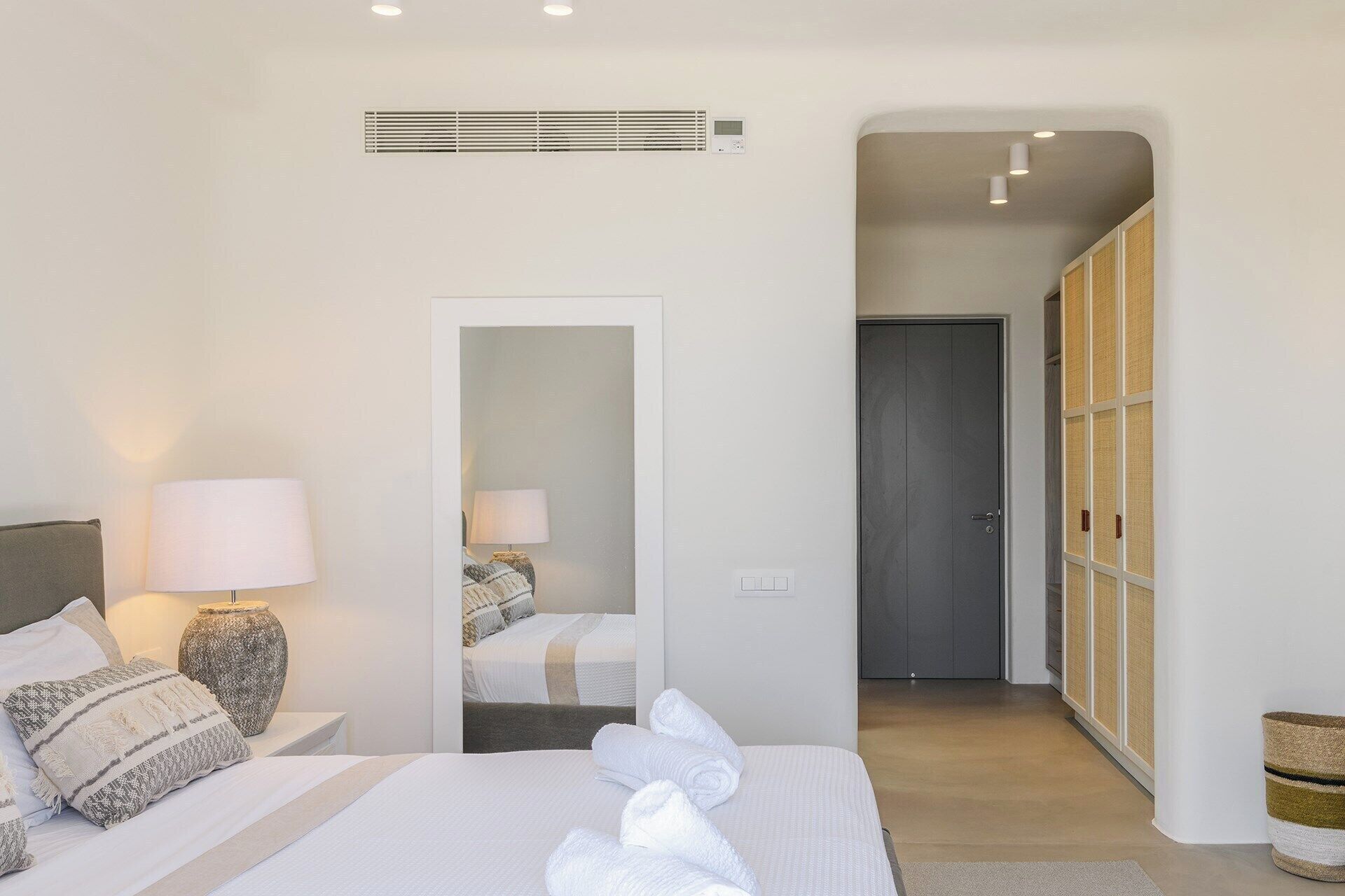 A bedroom with a bed, bedside lamps, a mirror, a wardrobe, and a door leading to another room.