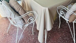 Uma área de refeições ao ar livre com uma mesa posta para quatro pessoas, rodeada por cadeiras de metal branco com almofadas estampadas.