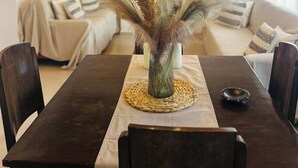 Uma área de refeições com uma mesa de madeira, cadeiras e um vaso com plantas secas. Um candelabro pendurado no teto, e um sofá visível ao fundo.