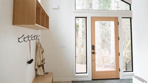 Un interior moderno con un banco de madera, luces colgantes, un perchero y un amplio ventanal con vistas a un bosque.