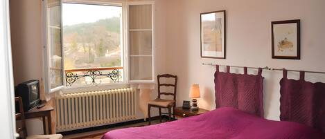 Ein Schlafzimmer mit Bett, Fenster mit Blick, Heizkörper, Stuhl, Tisch mit Lampe und Fernseher.