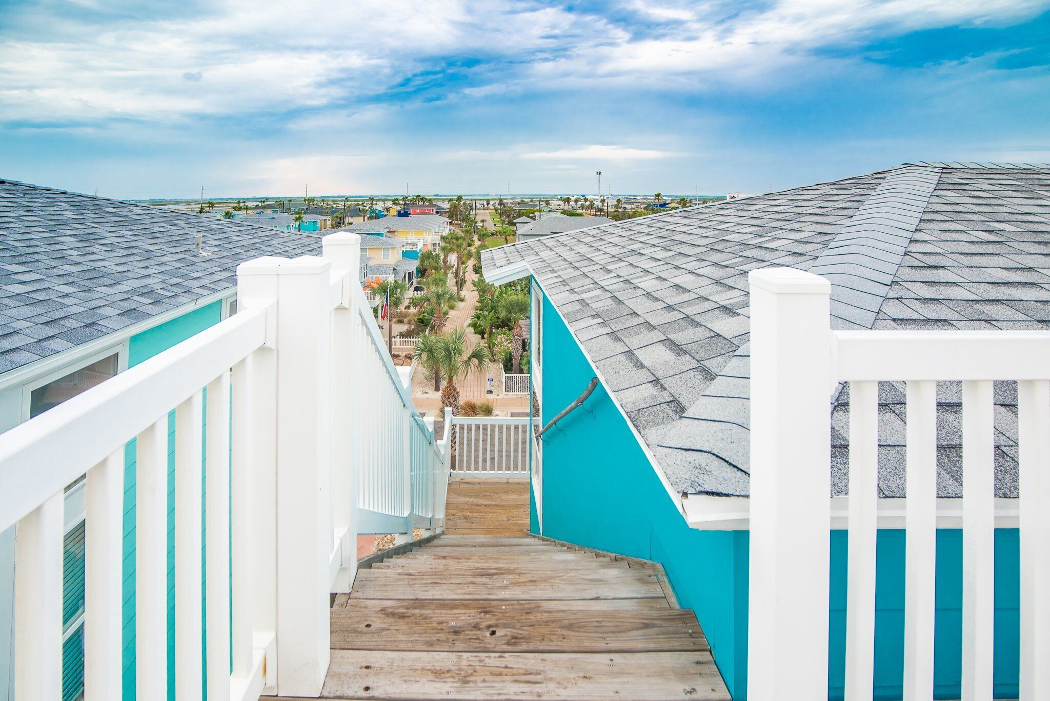 A wooden staircase leading to a balcony with turquoise walls and white railings, overlooking a beach and buildings.