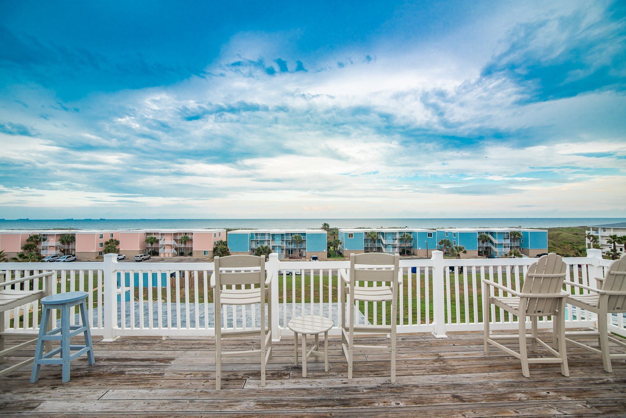 A wooden deck with white railings, overlooking a beach with a row of colorful buildings.