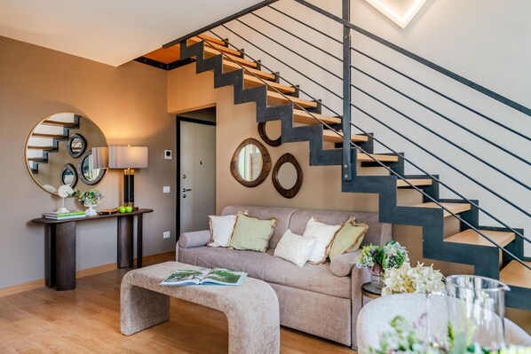 Una sala de estar moderna con una escalera, un espejo redondo, una consola con una lámpara y un sofá con cojines.