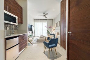 A modern kitchen with a microwave, sink, and wooden cabinets. There is a dining area with a round table and blue chairs. The room has a ceiling fan and a large wooden door. - Luxury Studio in Cabo San Lucas! Great Location, Pool, Gym & Paddle Court Access (Cabo San Lucas)