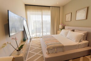 A modern bedroom with a large bed, a flat-screen TV mounted on the wall, and a window with sheer curtains. - Luxury Studio in Cabo San Lucas! Great Location, Pool, Gym & Paddle Court Access (Cabo San Lucas)