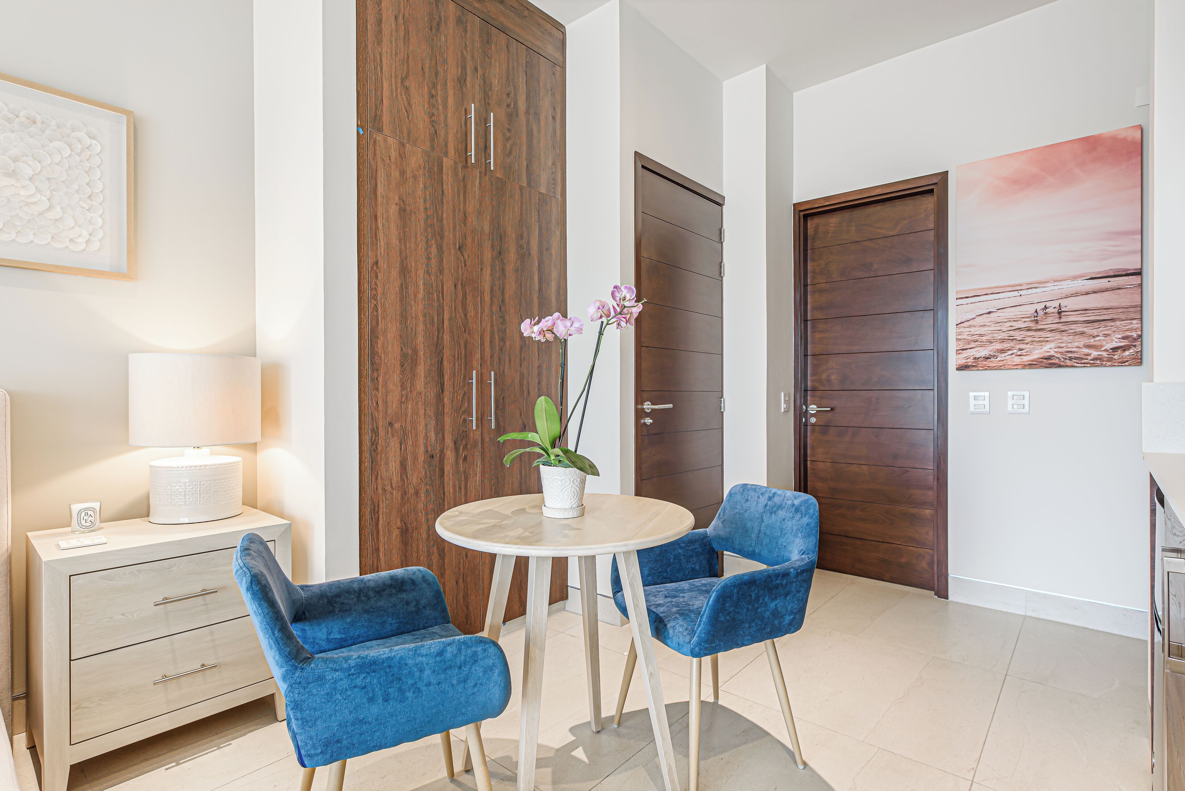 Een moderne kamer met een ronde tafel, twee blauwe stoelen, een witte lamp, een houten garderobe en een deur met een strandafbeelding.