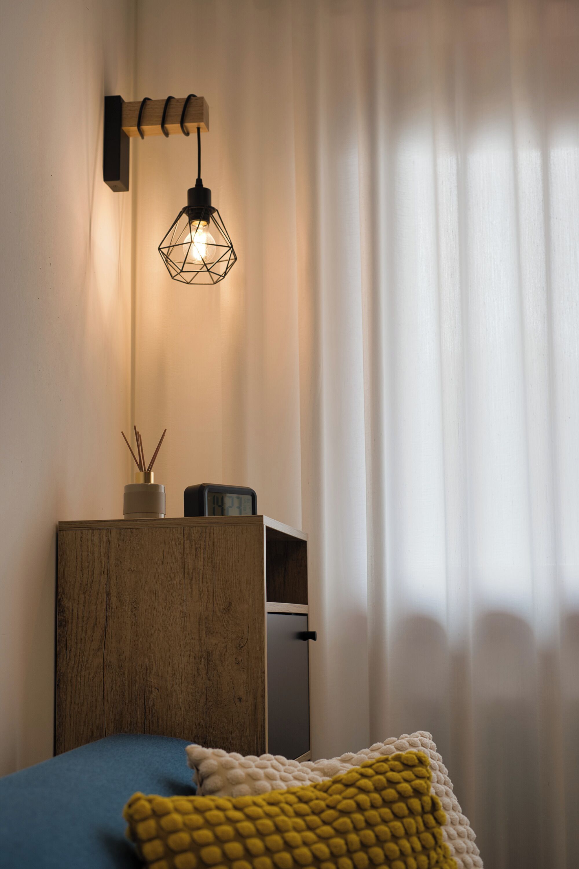 Quarto com cabeceira de madeira, mesa de cabeceira com relógio, luminária e janela com cortinas leves.