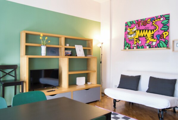 A room with a wooden desk, a white sofa, a TV, a bookshelf, and a colorful tiger painting on the wall.