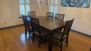 A dining room with a wooden table and chairs, a chandelier, and a large window with blinds. - Charming 3-bedroom country home (Manor)