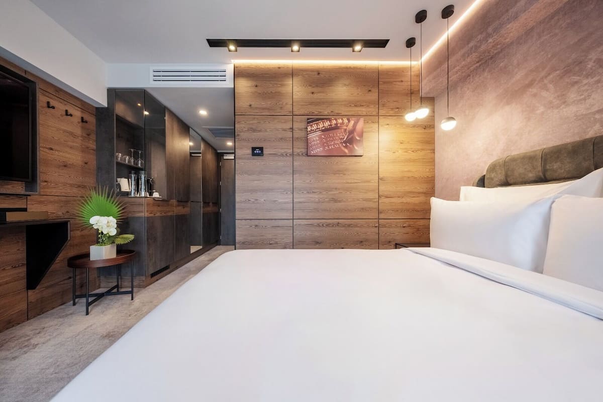 Moderni hotellihuone, jossa on suuri sänky, pieni sivupöytä, jossa on kasvi, puinen tehosteseinä ja seinälle kiinnitetty taideteos.
