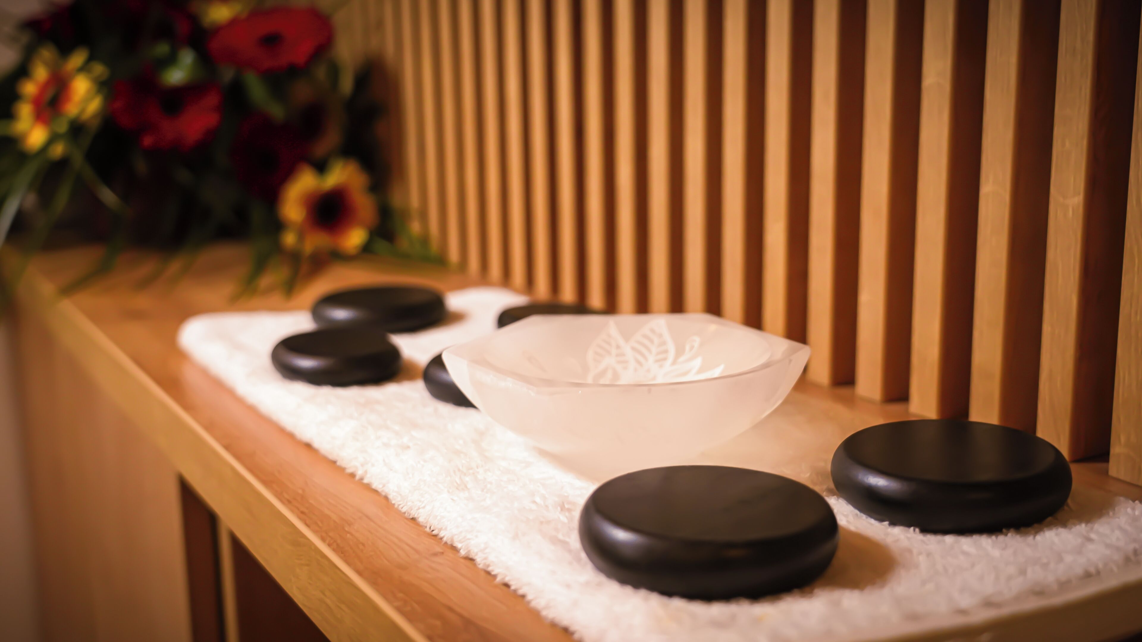 A wooden bench with a white towel, a clear glass bowl, and black stones, likely used for a spa treatment.