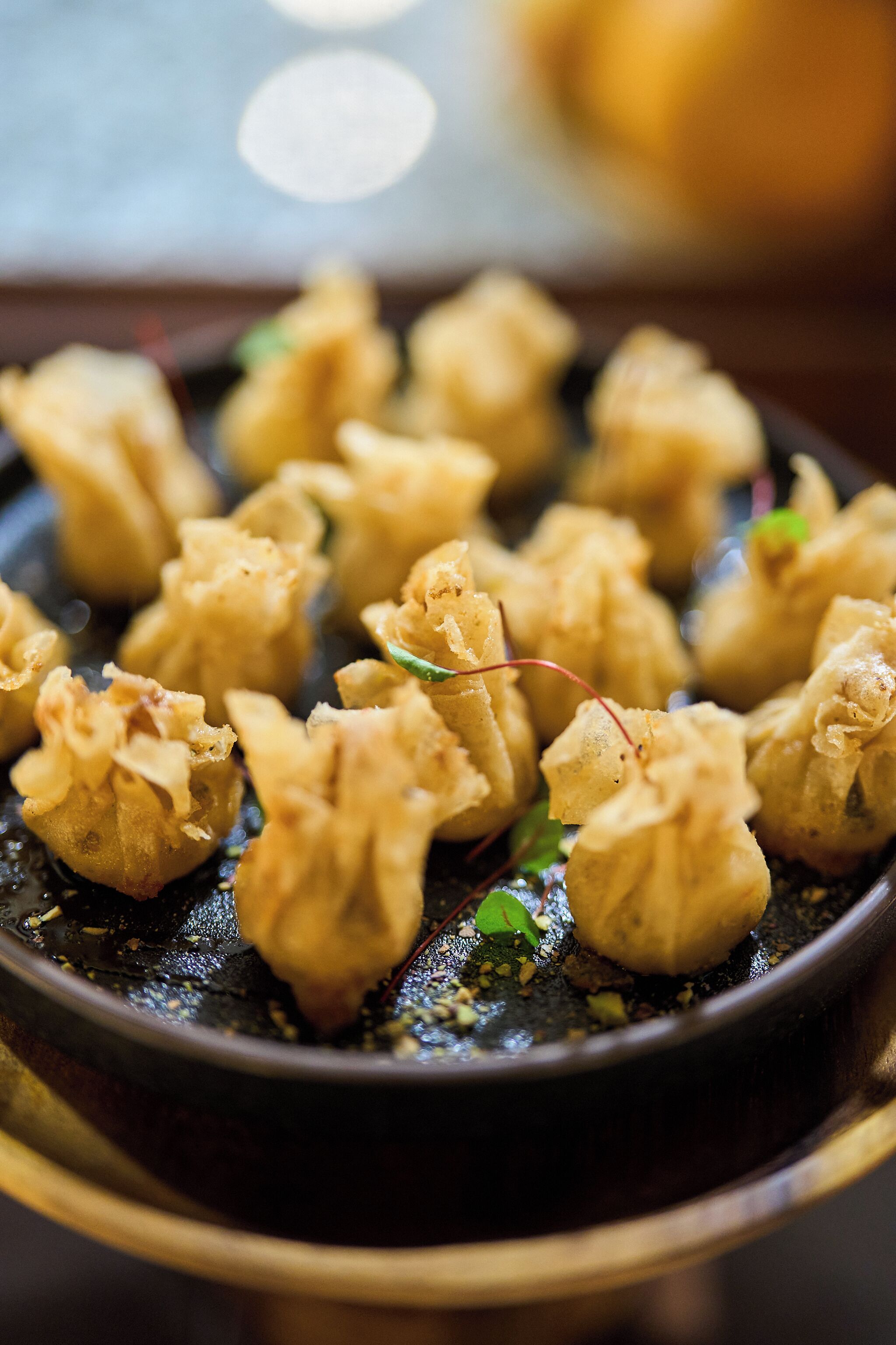 A close-up of a dish with dumplings, garnished with green herbs and a red leaf, served on a dark plate.