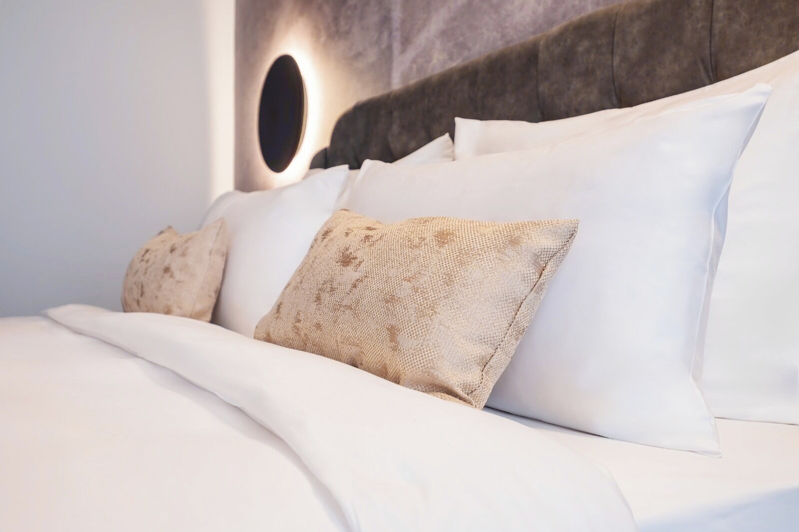 A neatly made bed with white linens and patterned pillows, a round wall-mounted light, and a textured headboard.
