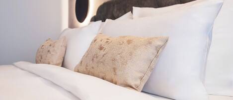 A neatly made bed with white linens and patterned pillows, a round wall-mounted light, and a textured headboard.