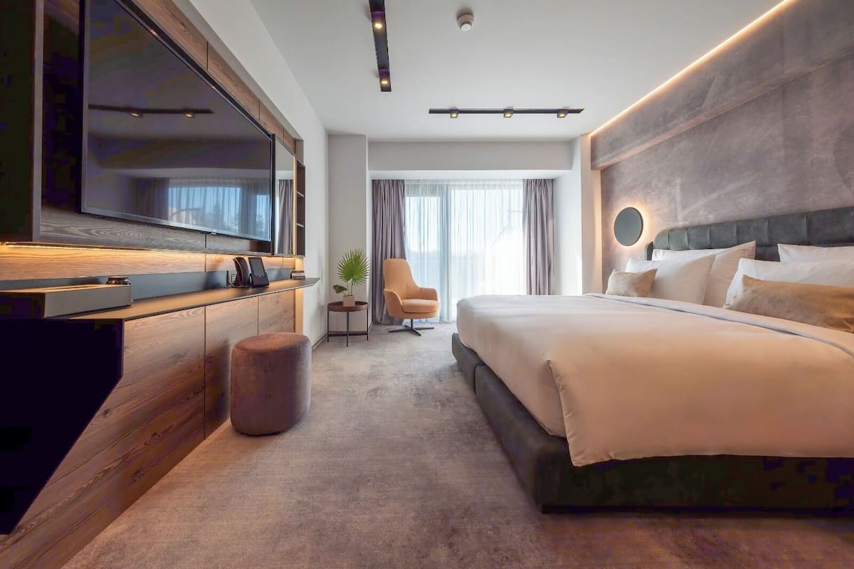 Moderni hotellihuone, jossa on suuri sänky, seinälle asennettu televisio, oleskelualue tuolilla sekä pieni sivupöytä.