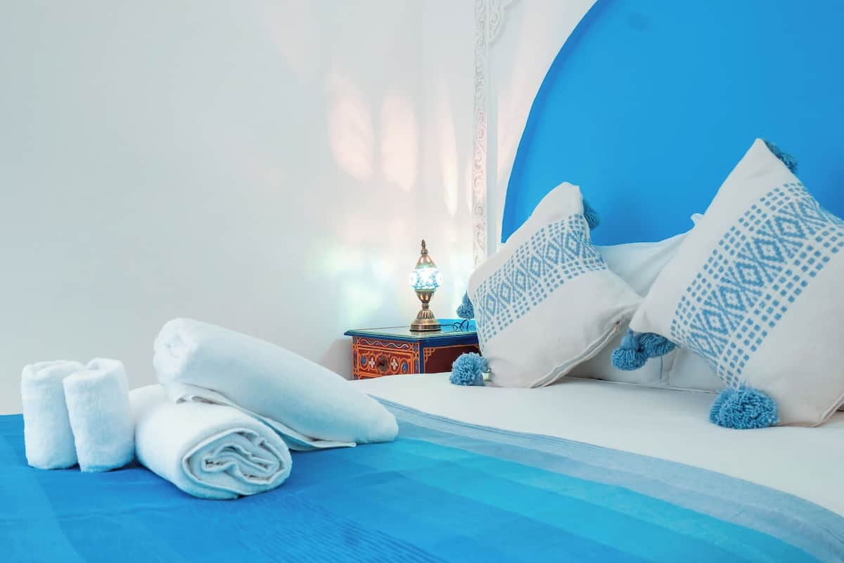 A neatly made bed with rolled white towels, a decorative lamp, and patterned pillows on a blue and white bedspread.