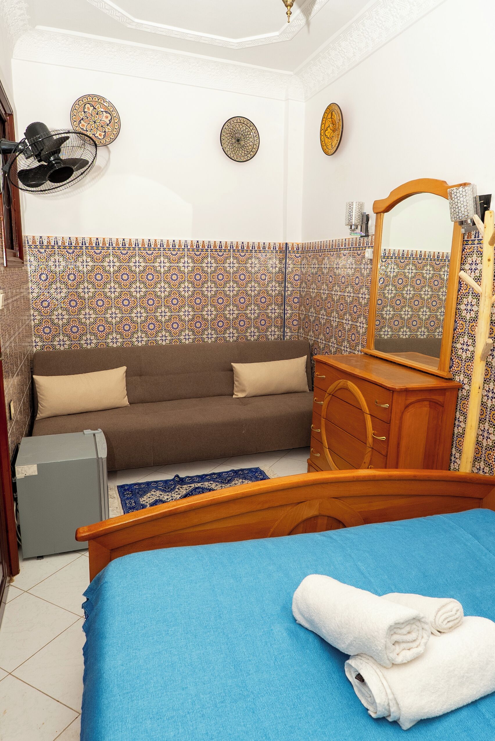 Un dormitorio con cama, cómoda de madera, sofá marrón, ventilador de pared y baldosas estampadas.