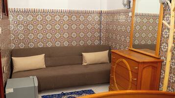 Un dormitorio con cama, cómoda de madera, sofá marrón, ventilador de pared y baldosas estampadas.